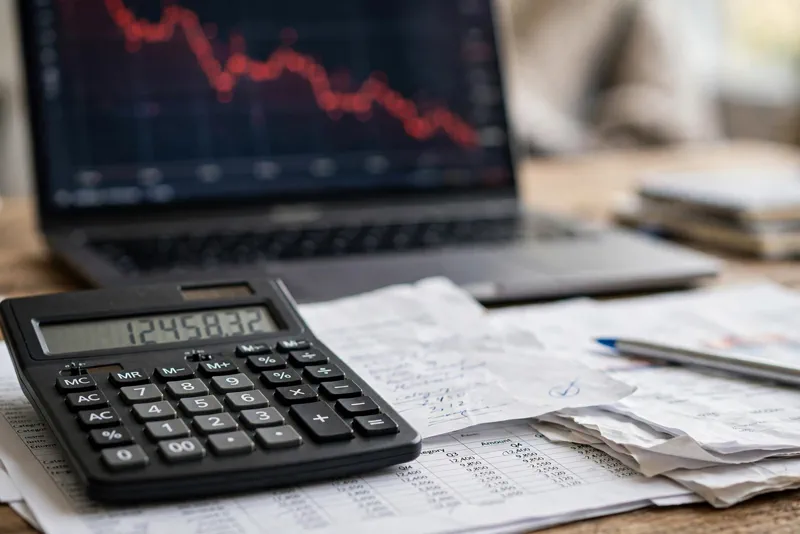 Calculator on financial documents with a declining chart on a laptop screen