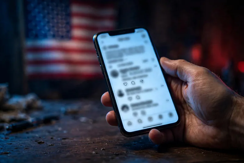 Hand holding a smartphone showing a Truth Social post, blurred American flag behind